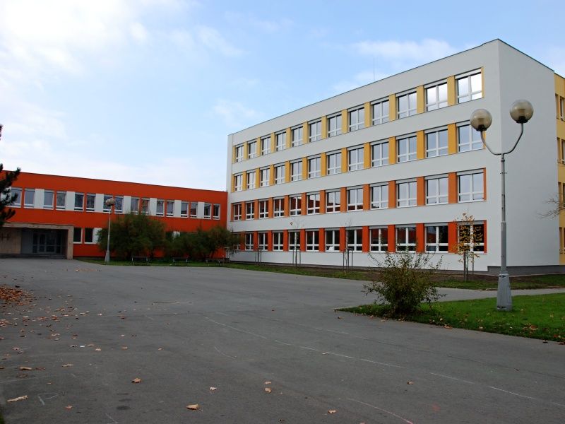 Moderní budova hradecké školy s bílými a oranžovými akcenty, velkými okny a třemi patry stojí uprostřed většinou prázdného dlážděného dvora s několika malými stromy a stožáry veřejného osvětlení a podporuje základní vzdělávání studentů.
