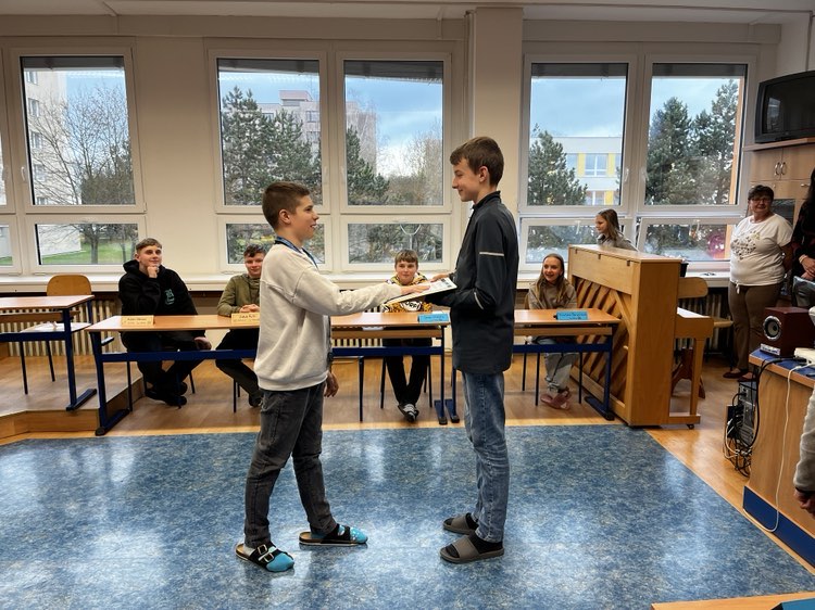 Dva chlapci stojí proti sobě ve třídě ZŠ Milady Horákové a jeden předává druhému vysvědčení. V pozadí přihlížejí sedící žáci a dospělí. Velká okna odhalují stromy a budovy venku.