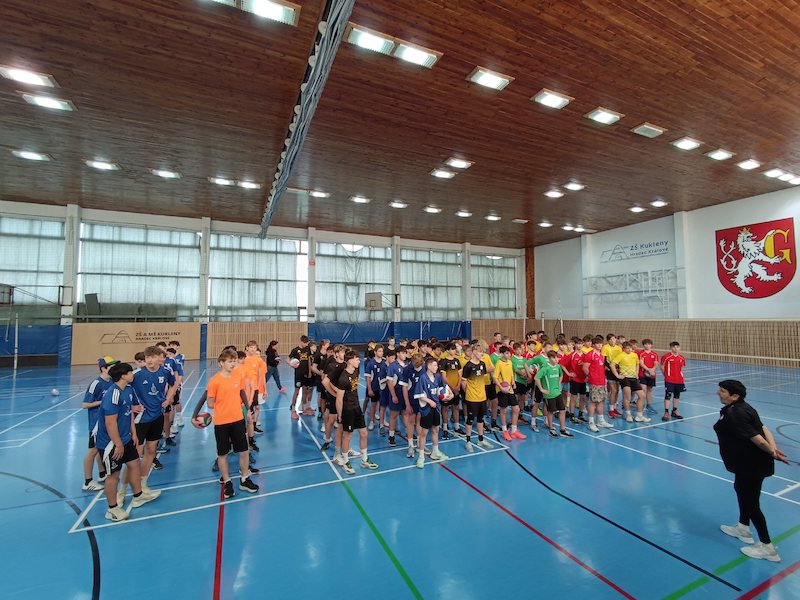 Velká skupina mladých sportovců ze Základní školy Hradec Králové v různobarevných dresech stojí v řadách na modrém krytém hřišti a naslouchá osobě v čele. Tělocvična má dřevěný strop a jasná, rovnoměrně rozmístěná stropní světla.