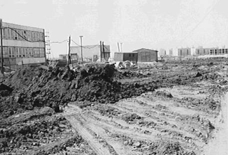 Černobílá fotografie zachycuje rozbahněné a rozježděné staveniště, na kterém bude stát budoucí Základní škola Hradec Králové, s hromadami zeminy, dočasnými budovami a částečně postavenými stavbami v pusté krajině.