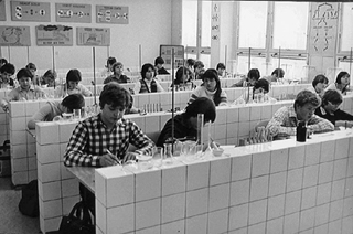 Černobílá fotografie žáků ZŠ Horákova, kteří pracují v laboratorních lavicích v učebně se zkumavkami a kádinkami; stěny lemují plakáty se schématy, zatímco velká okna zaplňují prostor přirozeným světlem.