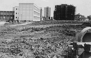 Černobílá fotografie staveniště u ZŠ Horákova, v popředí hlína a suť, v pozadí nedokončené budovy a výškové byty - pravděpodobně budoucí domy pro rodiny ze ZŠ Hradec Králové.